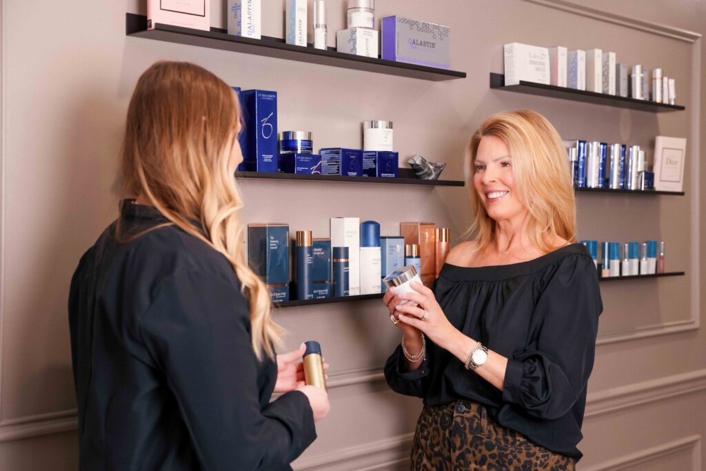 A female patient smiles in the clinic lobby, in front of a curated display of medical-grade skincare products. Her Look Lab provider is helping her pick the right ones to best support results from the best Hydrafacial in Denver.