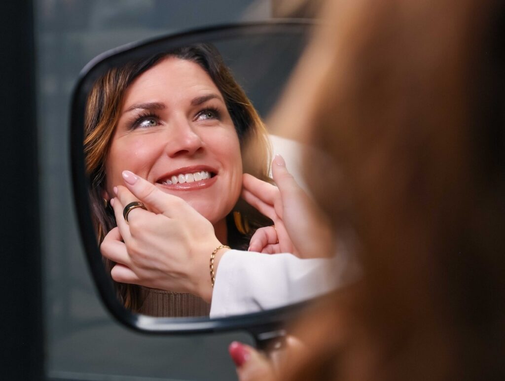 A female patient's smile reflected in a hand-held mirror as the provider shows where neurotoxin injections should be placed for the best Botox in Winter Park.